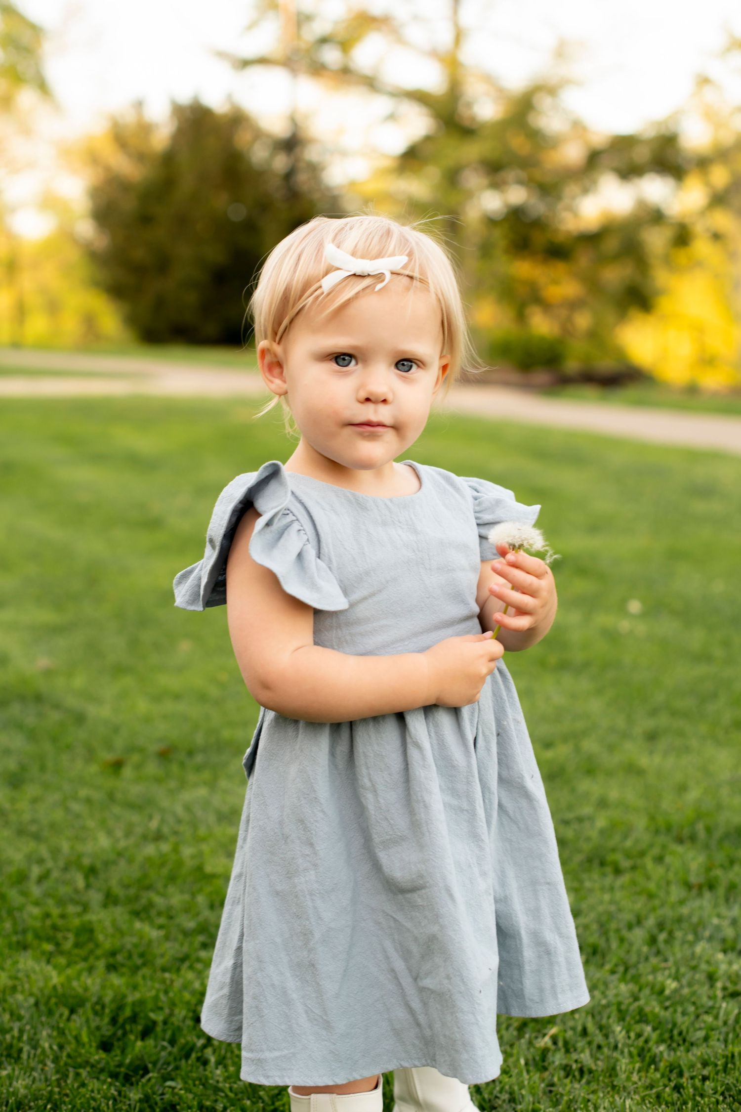 A small child in a light blue dress holds a flower while standing in a grassy area on a sunny day.