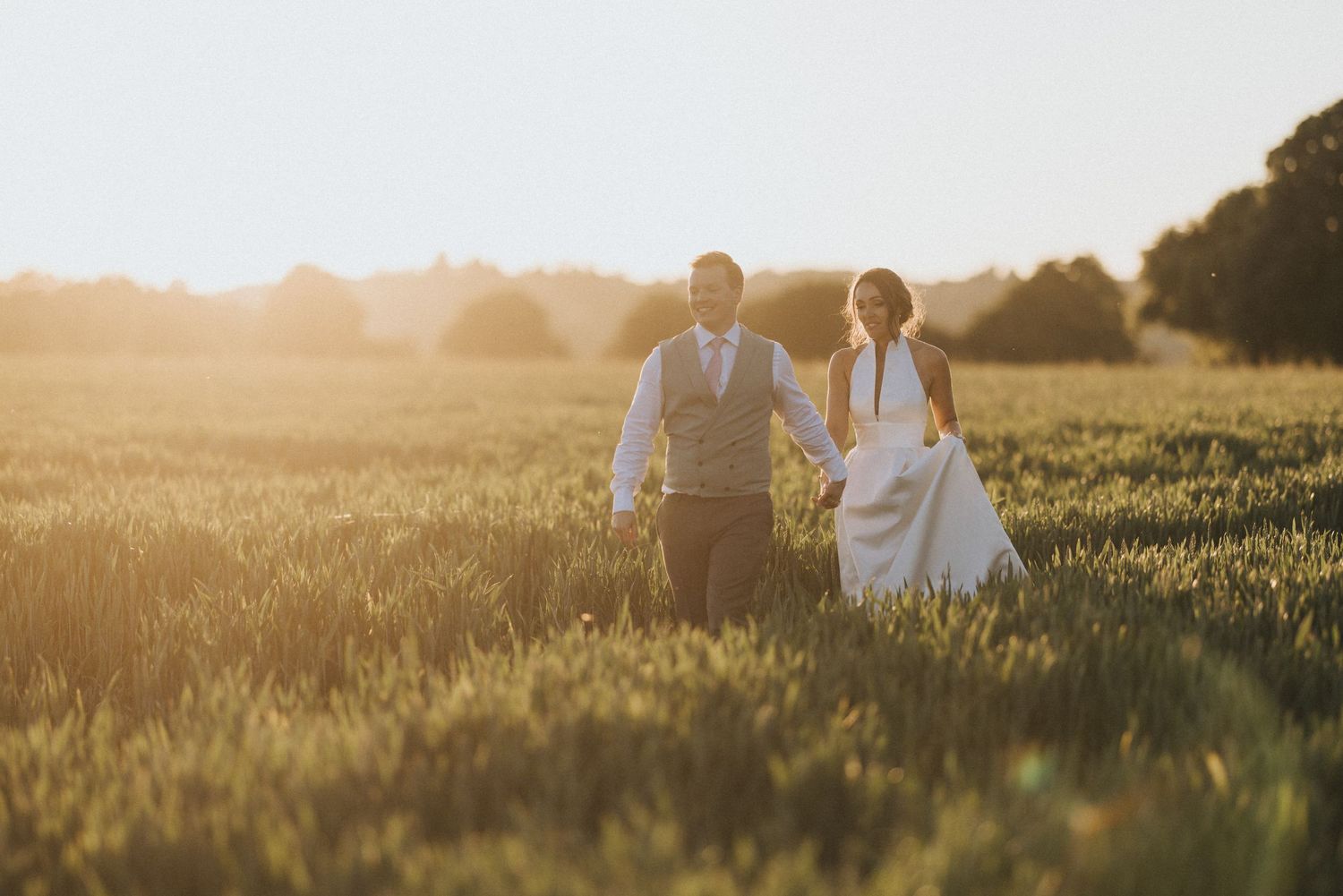 A couple in wedding attire walks through a golden sunlit meadow during sunset.