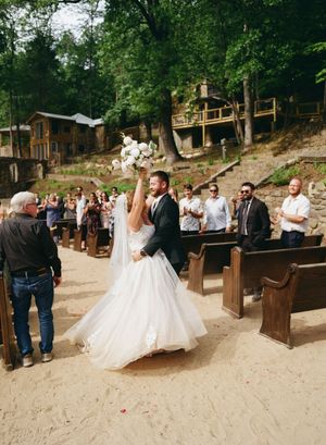 Bride raises bouquet joyfully as couple walks down sandy outdoor aisle lined with wooden pews amid cheering guests.