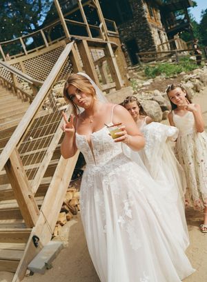 Bride in white lace gown poses on wooden stairs with bridesmaids at rustic outdoor wedding venue.