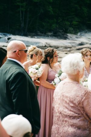 Bridesmaids in dusty rose gowns hold white bouquets during outdoor wedding ceremony near rocky landscape.
