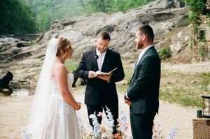 Outdoor wedding ceremony with bride in white gown, groom in dark suit, and officiant reading vows near rocky backdrop.