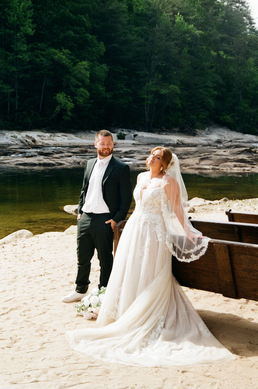 Bride in lace gown and veil poses with groom in dark suit on sandy riverbank surrounded by lush forest.
