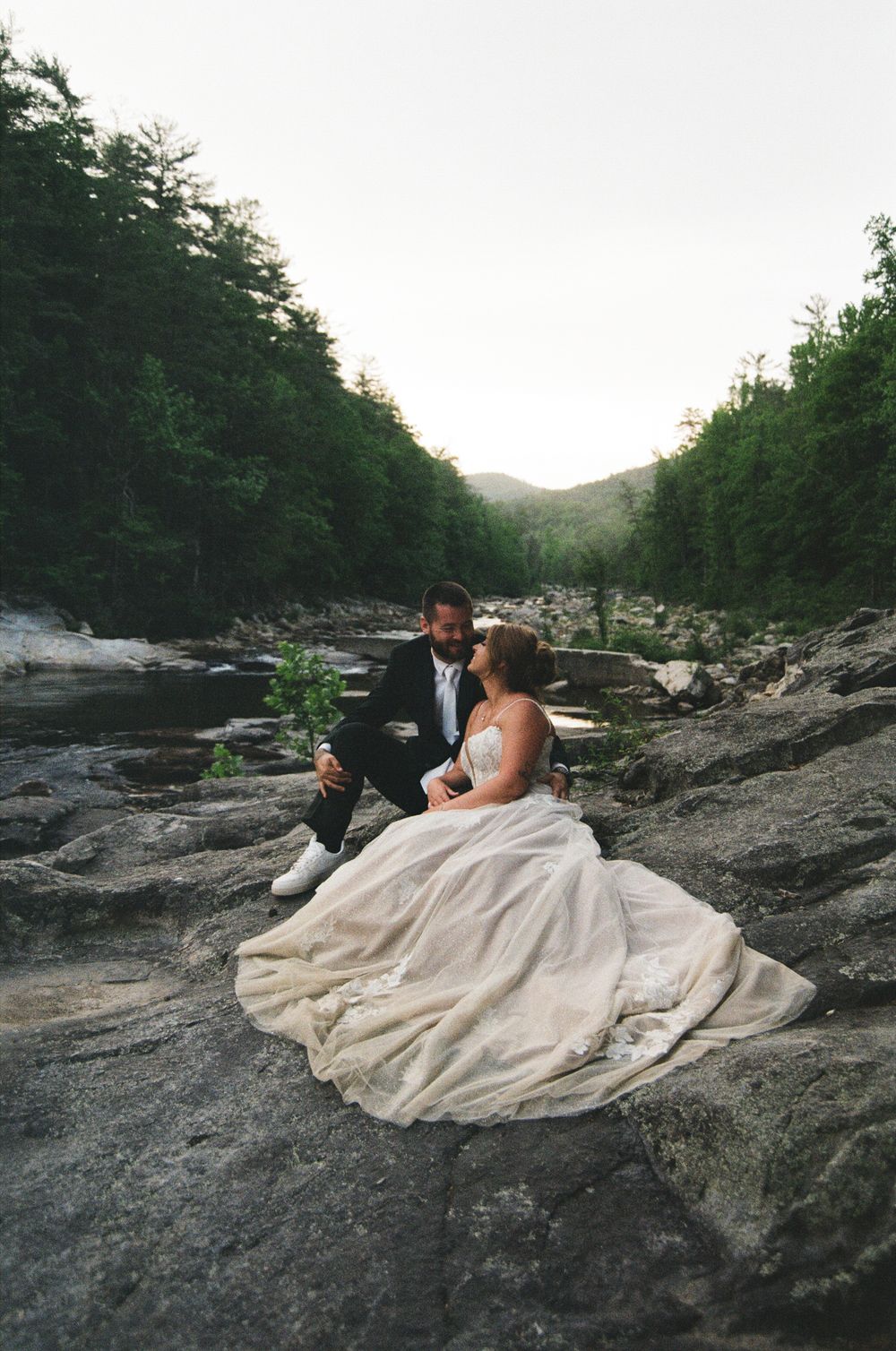 Bride and groom in wedding attire sitting on rocky riverbank surrounded by lush green forest at dusk.