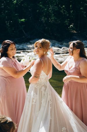 Bride in lace gown with bridesmaids in pink dresses helping her prepare outdoors near a creek.