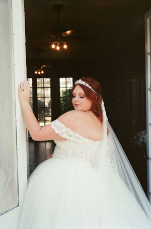 Bride in off-shoulder lace ballgown with tiara and veil poses in doorway, looking down elegantly.