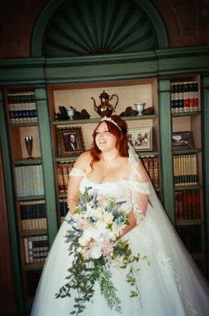 Smiling bride in white off-shoulder gown holding cascading floral bouquet, standing before a bookshelf in an elegant library.