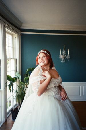 Bride in a white ball gown with lace details and tiara smiles in a teal-walled room with natural window light.
