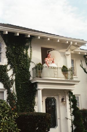 Bride in white gown holding parasol on ivy-covered mansion balcony with ornate railing on a cloudy day.