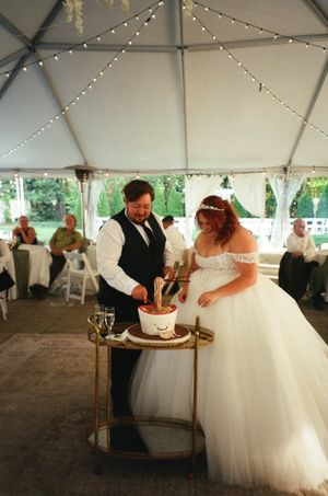 Bride and groom cutting wedding cake under a white tent with string lights at an outdoor reception.