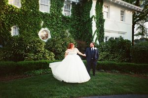 Bride in white gown twirls with groom in navy suit outside ivy-covered estate on their wedding day.