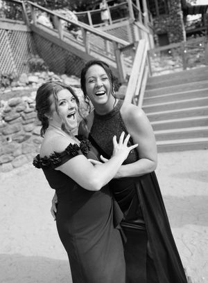 Two women in elegant black gowns laughing and embracing outdoors near stone steps in a black and white photo.