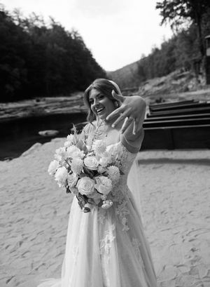 Smiling bride in lace wedding gown holding white floral bouquet and showing ring on sandy riverbank.