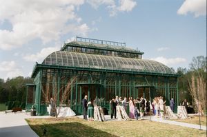 Panoramic view showing six connected glass greenhouse structures with people gathering outside in a garden setting.