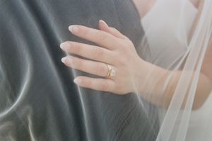 A delicate hand wearing an engagement ring rests against a grey fabric, with soft white veil material visible.