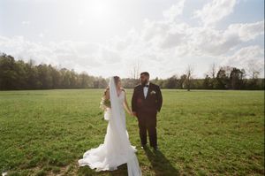 A rustic country wedding scene with a couple holding hands in a vast green meadow during golden hour.