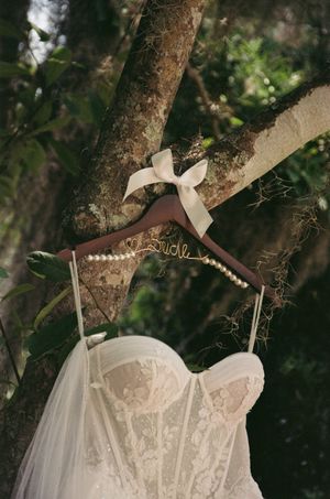 White lace wedding dress with corset bodice hanging on wooden hanger from tree branch in natural outdoor setting.