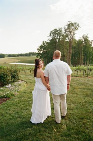 Bride and groom hold hands facing a scenic vineyard and rolling green hills at golden hour.