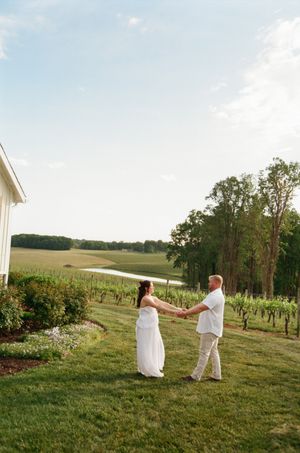 Couple in white holding hands in a vineyard with a pond and trees in the background on a sunny day.