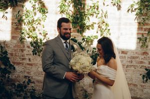 Bride and groom smiling together, holding white floral bouquet, brick wall with hanging greenery backdrop.