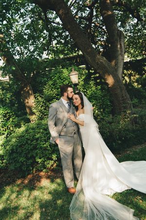 Romantic bride and groom embracing under lush green trees with veil flowing in the breeze.