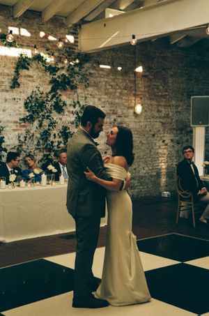 Bride and groom share their first dance on a checkered floor in a rustic brick venue with ivy and Edison bulb lighting.