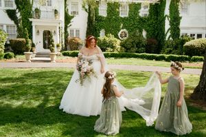 Bride in white gown with two flower girls in sage green dresses holding veil on lush garden lawn.