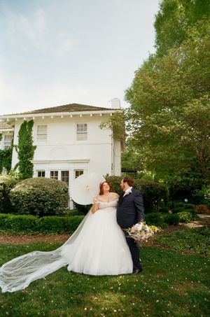 Bride in white gown holding parasol stands with groom in navy suit on lush garden lawn before elegant white estate.