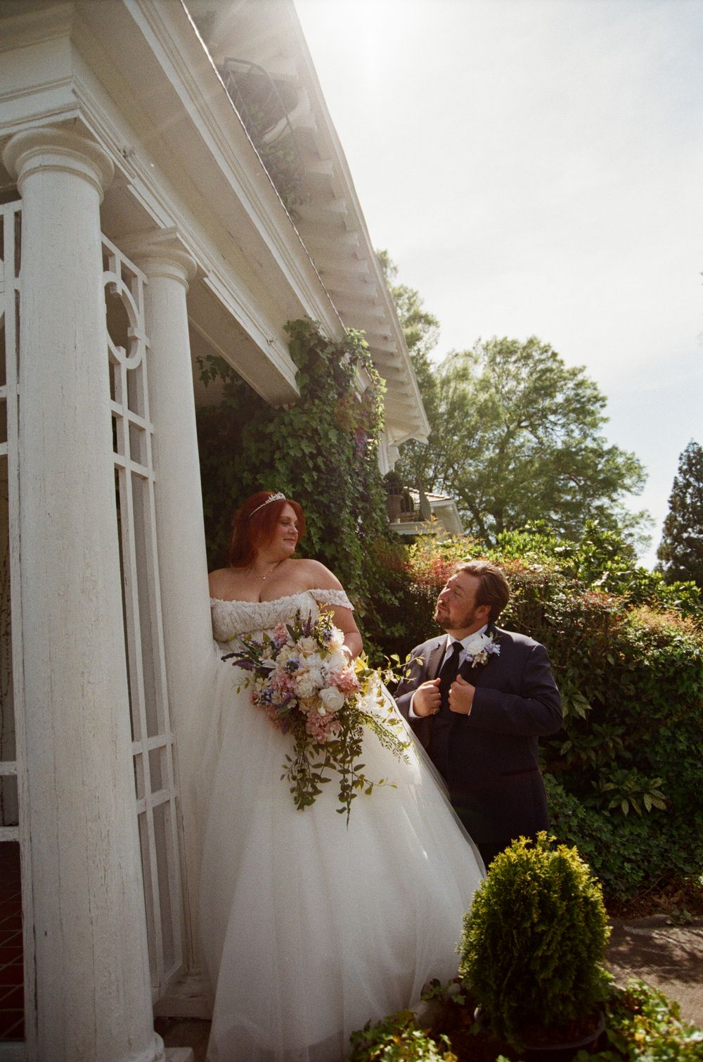Bride in off-shoulder gown holding floral bouquet stands with groom in navy suit by white columns of elegant venue.