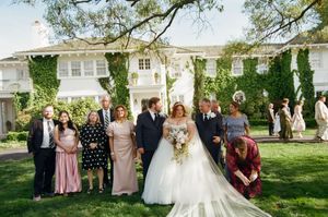 Wedding party gathers on lush lawn before ivy-covered white manor house during outdoor ceremony.