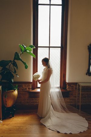 Bride in white gown and veil holding bouquet stands by tall window with natural light in elegant venue.