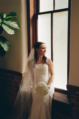 Bride in white gown and veil holding white bouquet standing by large window in elegant venue.