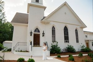 Bride in white gown posing outside a charming white chapel church with arched windows and wooden door.