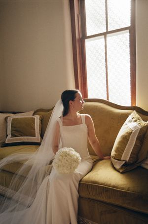 Bride in white gown and veil sits on gold velvet sofa holding white bouquet, gazing out sunlit window.