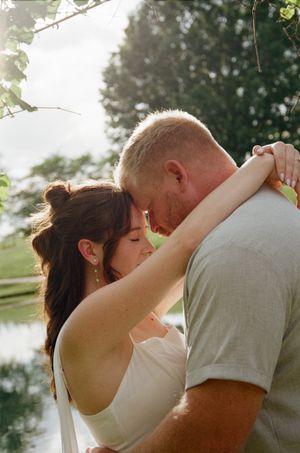 A couple shares a tender forehead touch embrace outdoors near a pond, surrounded by lush green trees in soft natural light.