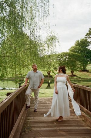 Bride in white dress and groom in gray shirt walk on wooden bridge surrounded by weeping willow trees and pond.