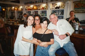 Four friends pose together smiling inside a cozy bar with shelves of bottles in the background.