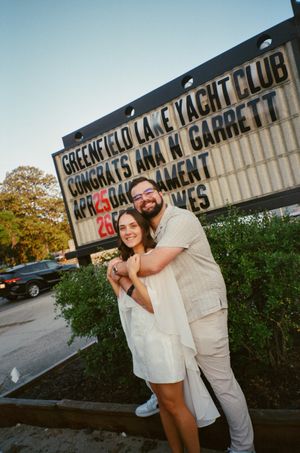 Couple embracing and smiling in front of Greenfield Lake Yacht Club congratulations sign for Ana and Garrett.