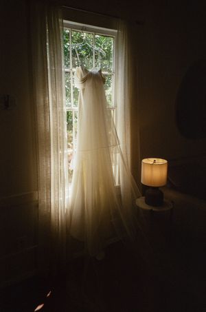 Elegant white wedding dress hanging in window with warm lamp glow in dark room.
