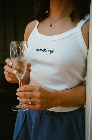 Woman wearing a white tank top embroidered with garrett's wife, holding a champagne flute, with a ring and necklace.