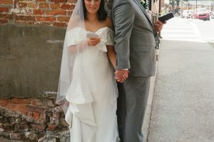 A bride in a white gown and veil holds hands with a groom in a gray suit outside a brick building.