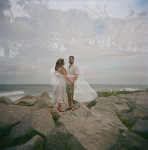 A couple walks along rocky coastal cliffs as waves crash against the shore on a cloudy day.