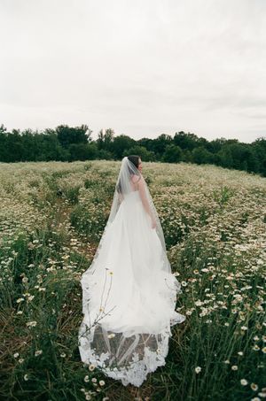 A flowing white wedding dress and veil stands ethereally among a field of wild daisies and summer flowers.