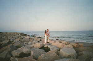 A couple walks hand in hand on a rocky beach jetty at sunset with ocean waves in the background.