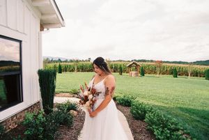 A bride in a white gown holds a bouquet while standing beside a building with green farmland in the background.