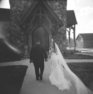 A stunning black and white photo of a wedding couple walking down a church path with a flowing white veil trailing behind.
