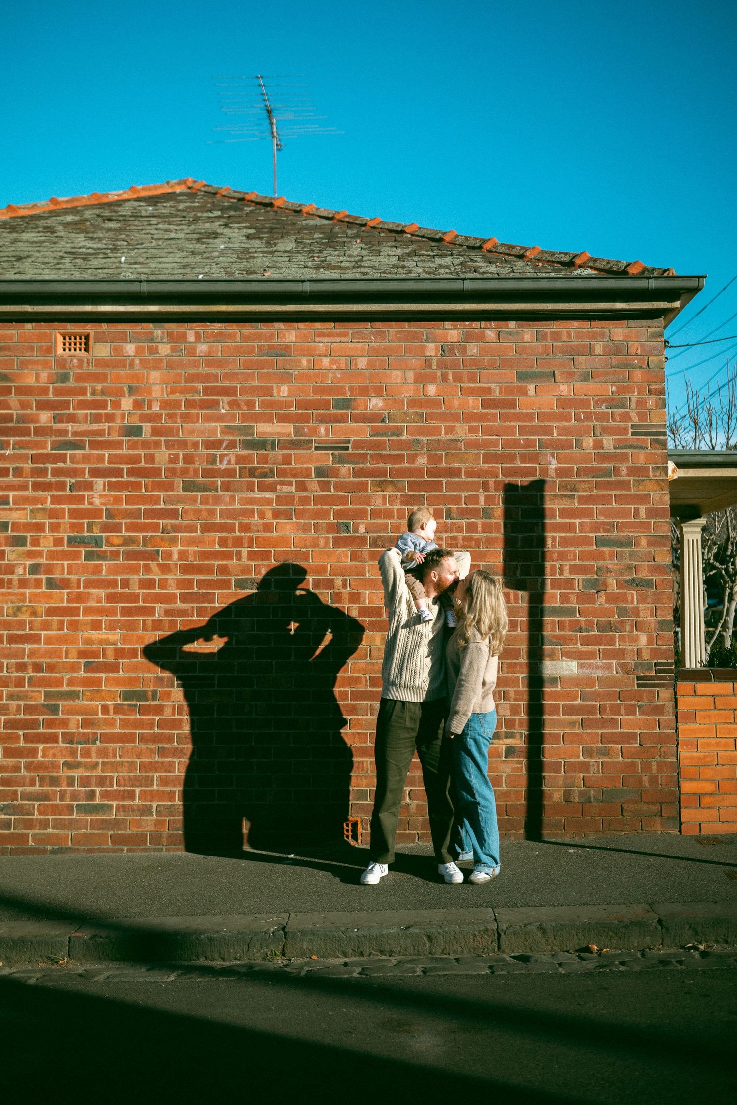 A young family stand against a red brick building wall as a shadow is cast on the surface during a family photoshoot in Richmond.