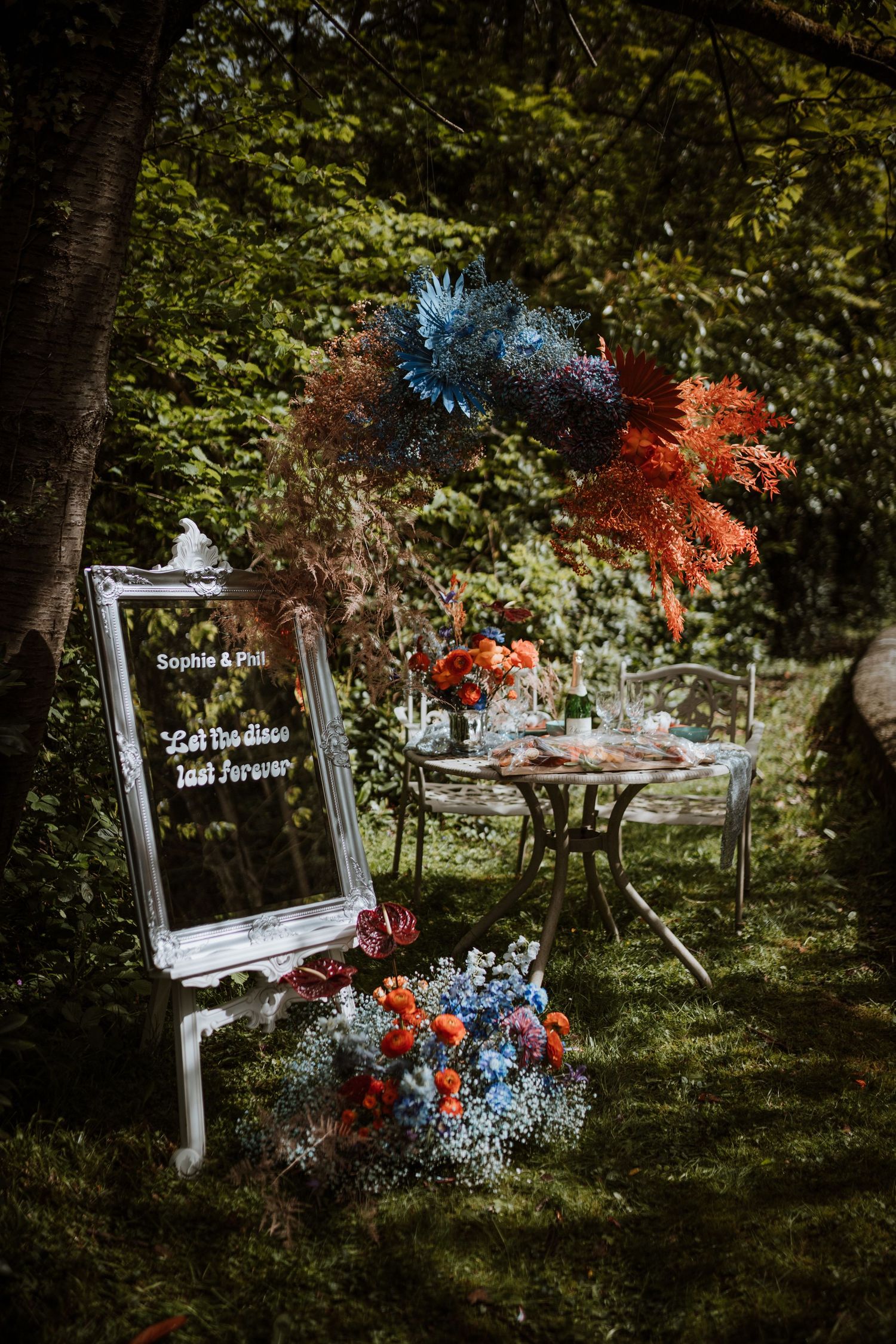 A romantic garden wedding setup with a mirror sign and blue and red floral arrangements in a woodland setting.