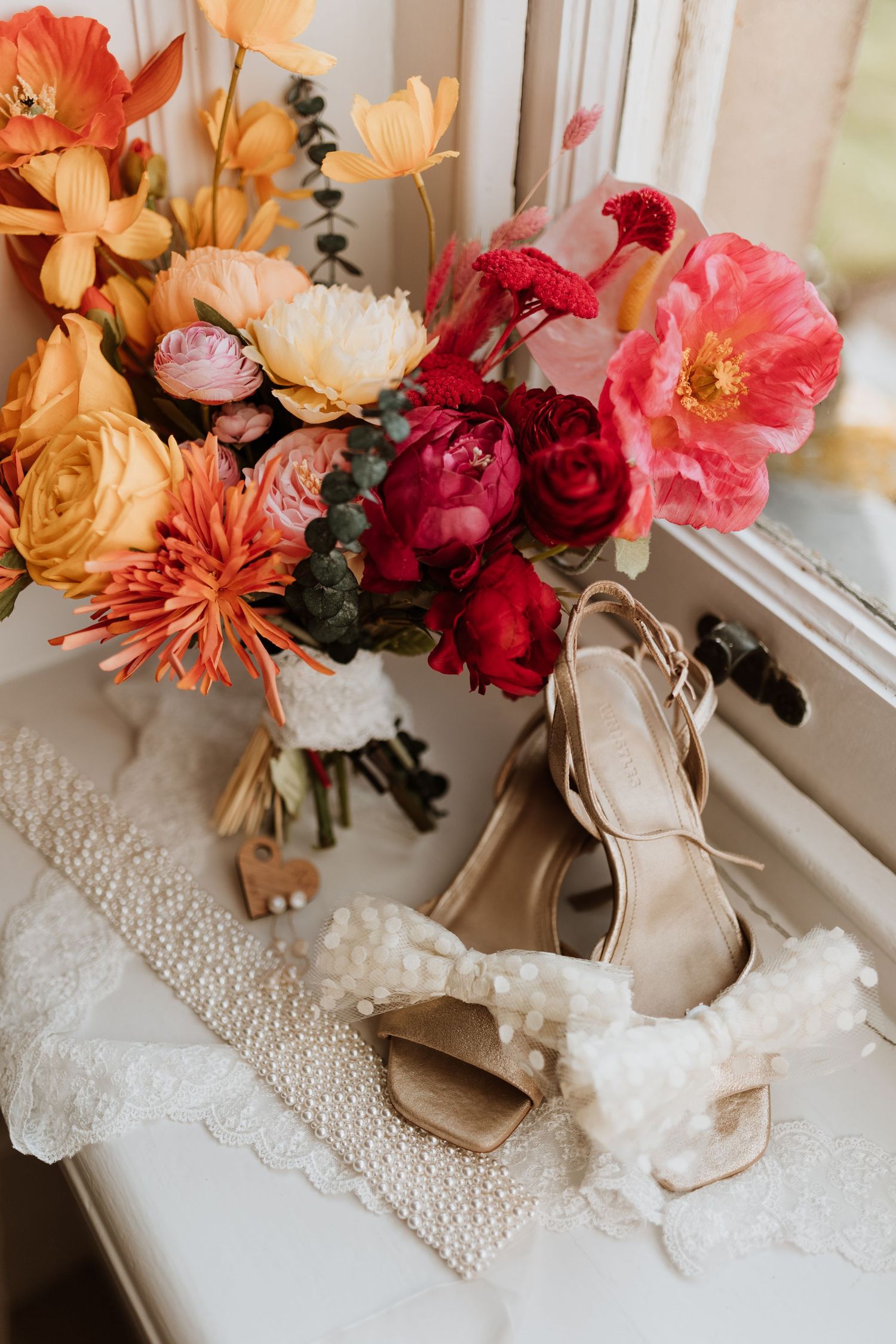 Elegant bridal shoes next to vibrant coral and burgundy floral arrangement with roses and poppies on windowsill.