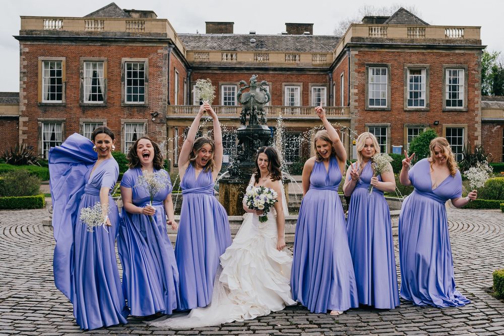 Bridal party in periwinkle blue bridesmaid dresses pose together outside historic brick building on wedding day.